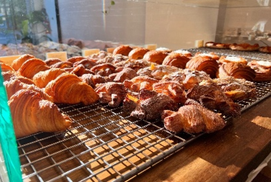 Metal cooling racks of cooked pastries including croissants, and other assorted pastries, on a wooden shop counter in a bakery.