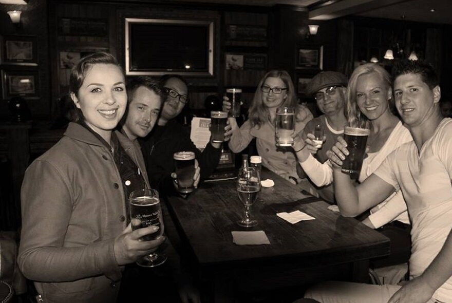 A group of people seated around a table in a dark pub, holding pints of beer.