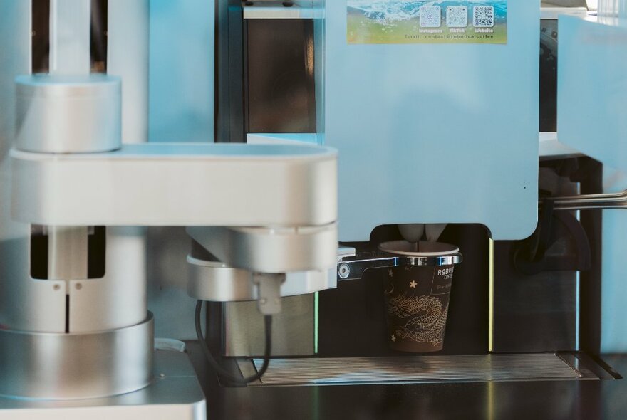 A robot barista placing a brown takeaway coffee cup under a blue machine.