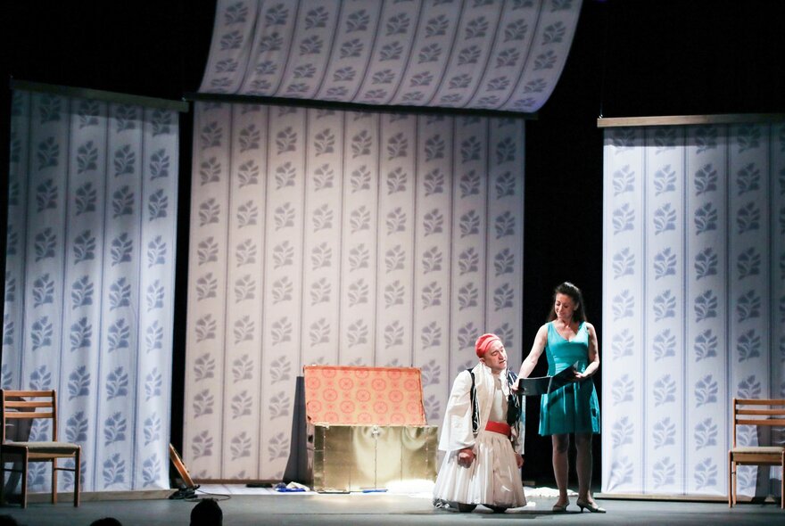Two actors conversing and interacting on a theatre stage, with a background of patterned wallpaper.