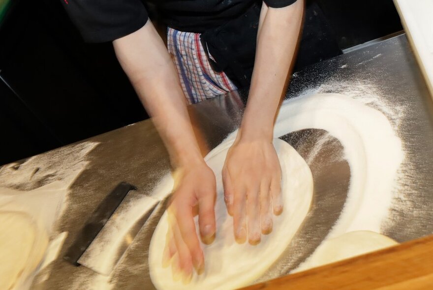 A pair of hands shaping dough on a floured table into a round pizza.