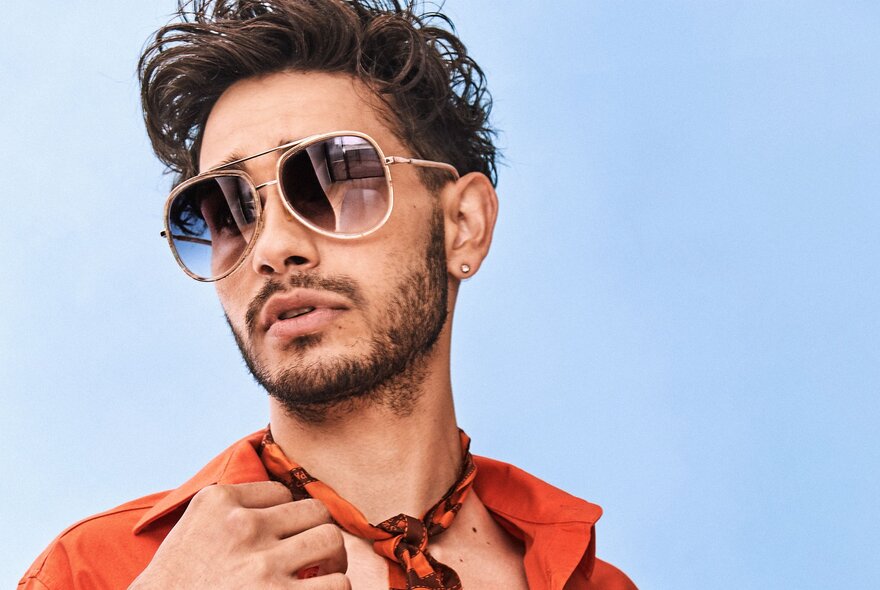 A man in purple-fade sunglasses, wearing an open orange shirt and an orange bandana, against a light blue background.