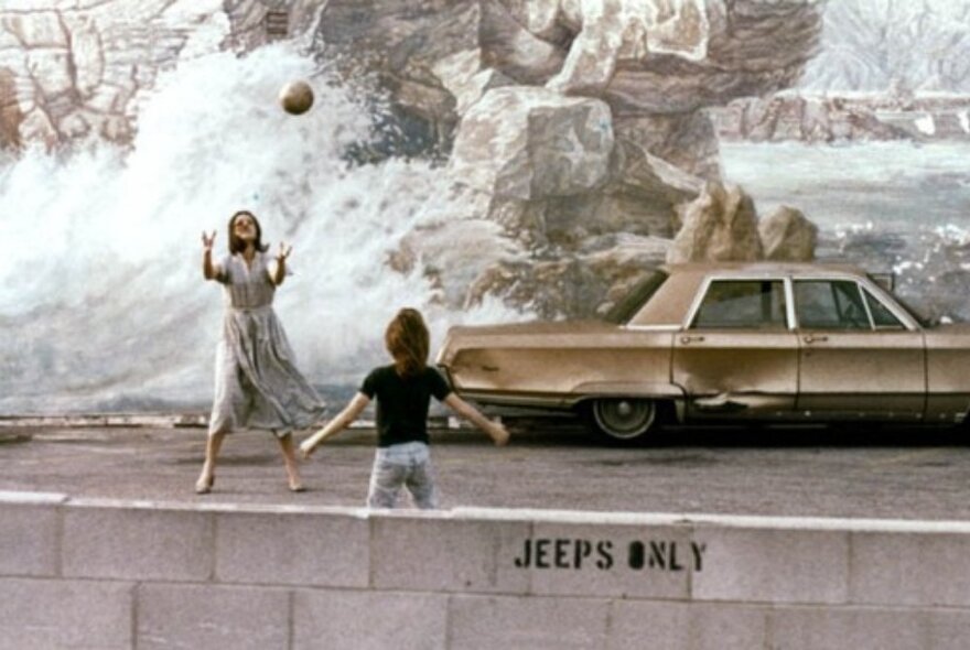 A still from the film, Mur Murs, depicting a mother and son playing catch in front of a giant mural painting of waves crashing against rocks, with a beat-up old chevy in front of it.