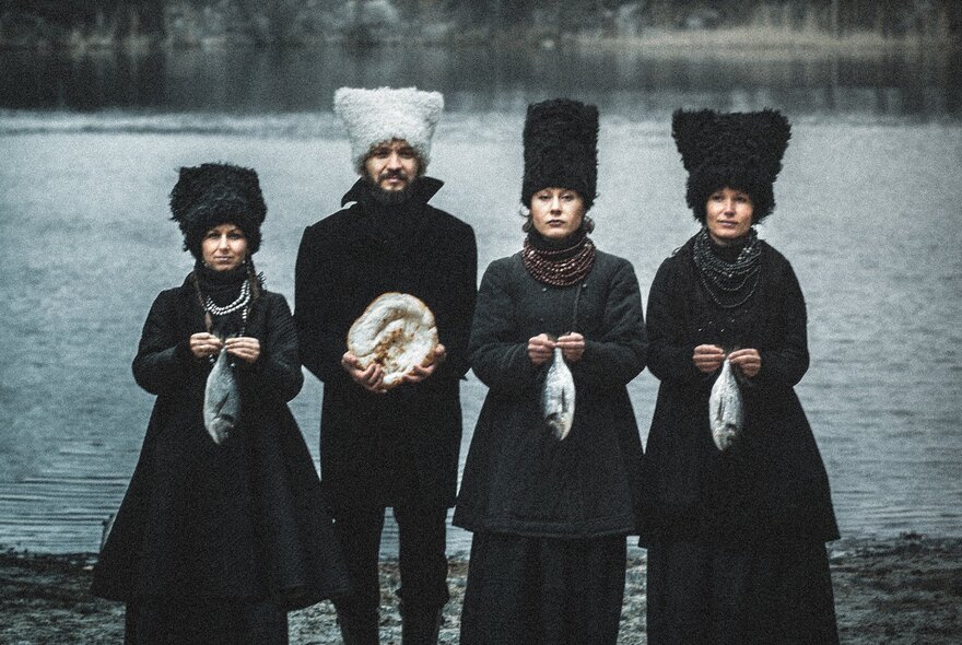 Four members of the group DakhaBrakha in oversized wooly hats and coats beside a lake, each holding a fish.