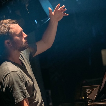 A dimly lit room with a man in a khaki t-shirt conducting an orchestra, hand raised. 