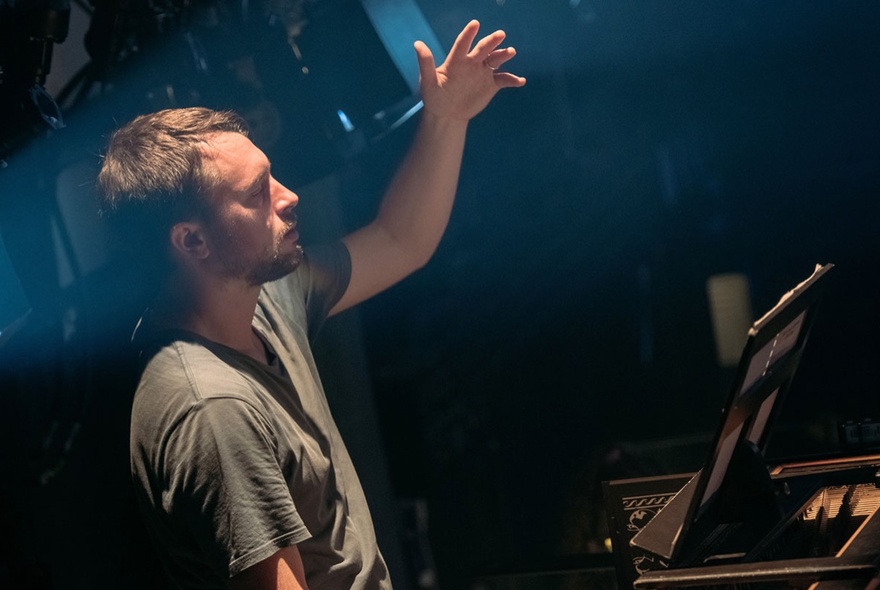 A dimly lit room with a man in a khaki t-shirt conducting an orchestra, hand raised. 