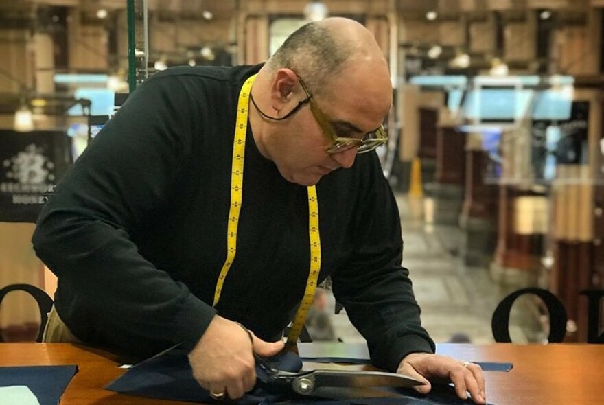 Tailor at work with large scissors on bench, and yellow tape measure around his neck.