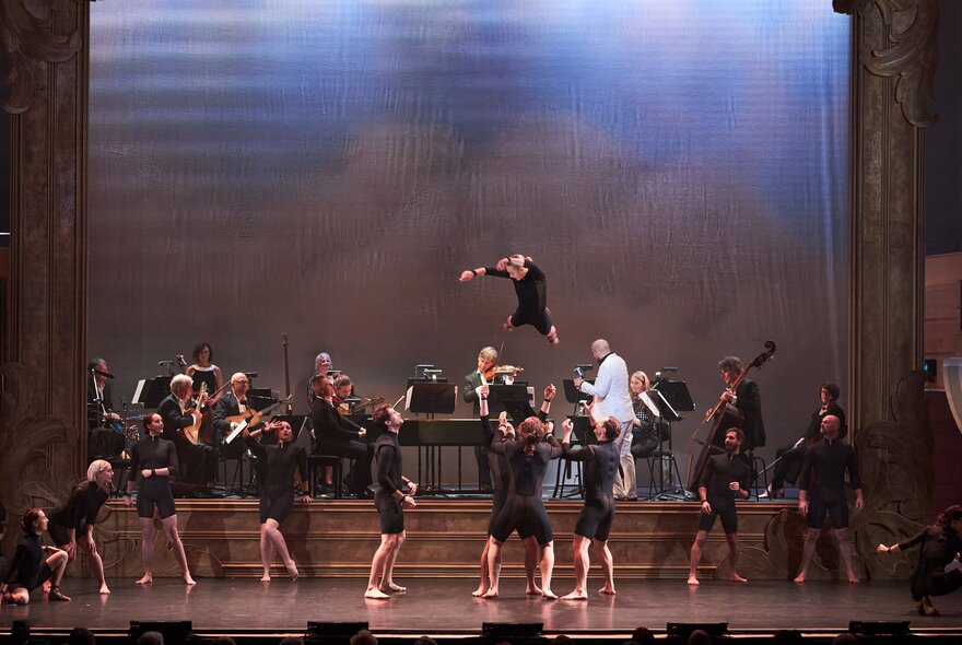 A group of circus performers on stage performing acrobatics, while an orchestra seated behind them plays music.