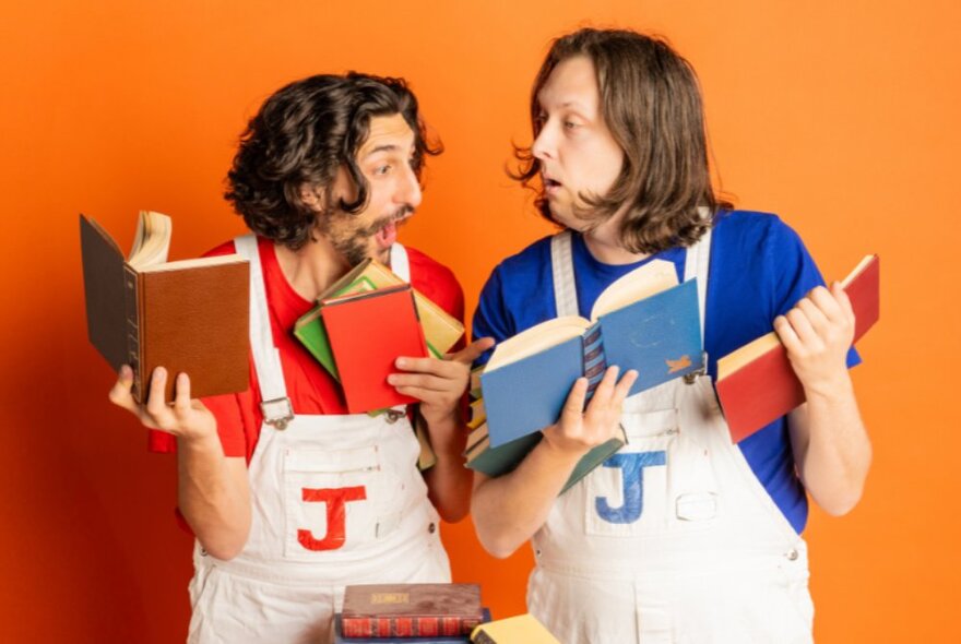 Two comedians in white overalls with bemused and silly expressions on their faces, holding multiple books in their hands and arms, standing against an orange background.