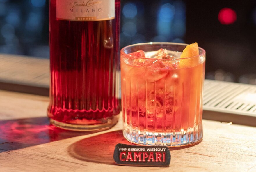 Tight shot to a Negroni with a Campari bottle in the background and small Campari label near the base of the glass. 