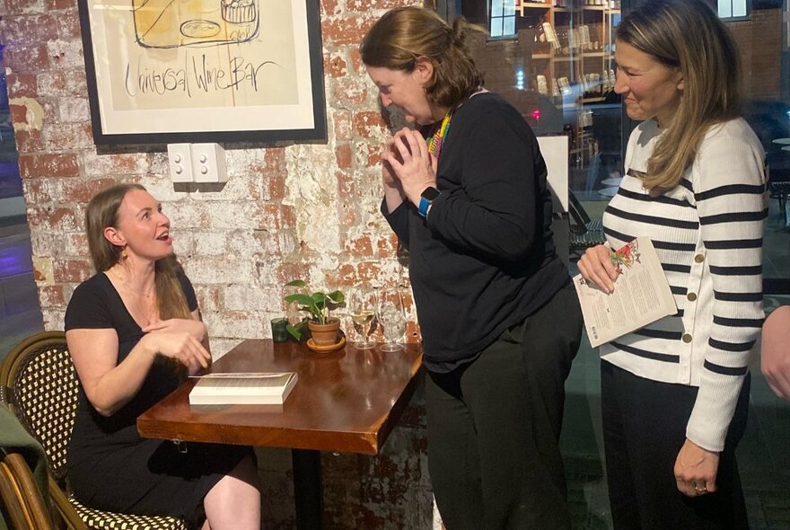 An author signing a book at a table, speaking to people queuing with books for signing.