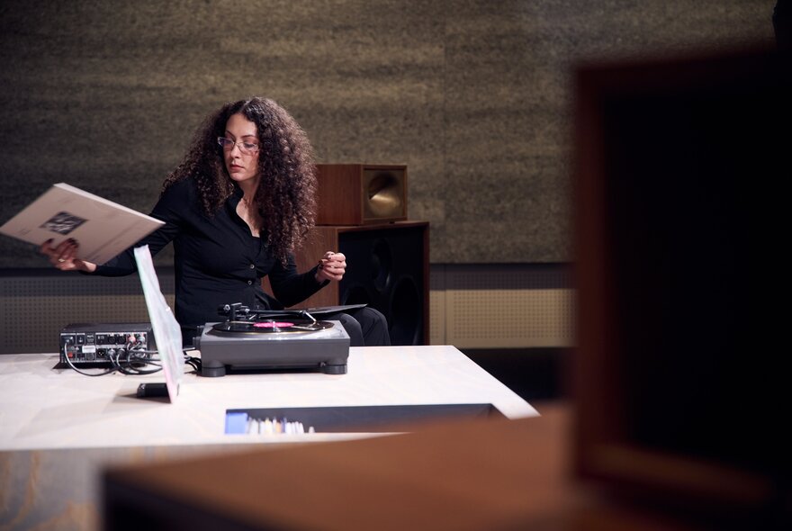 A seated person holding a vinyl record as if about to play it on a turntable on a low table in front of them. 