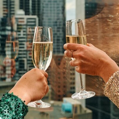 Two hands raising two flute glasses of sparkling wine against a backdrop of city skyscrapers seen though a window.
