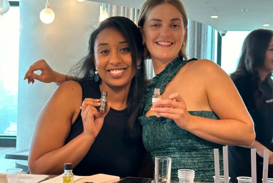 Two smiling women at a perfume workshop holding small vials and huddled together. 