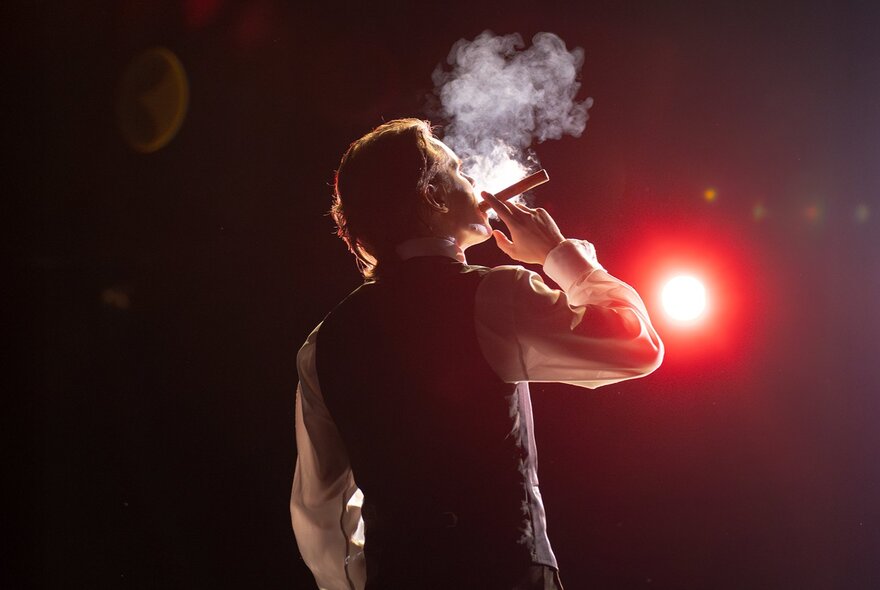 Man smoking a cigar viewed from behind with smoke illuminated by red light in dark room.
