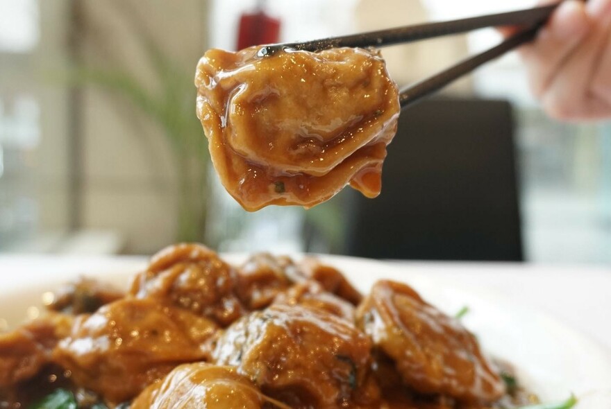 Dish of glossy tofu with one being picked up with black chopsticks. 