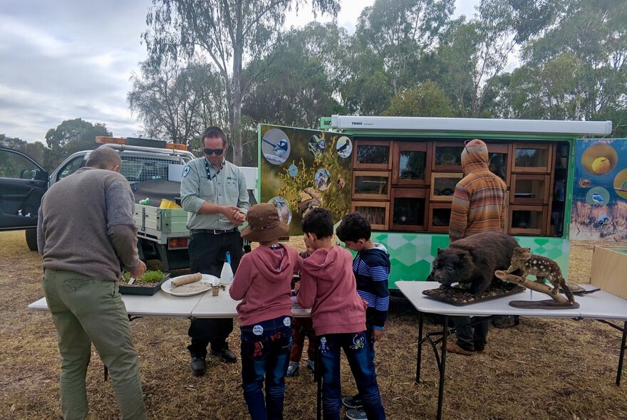 A man with two trestle tables showing objects to kids in a park environment, an open van with shelving and boxes behind him. 