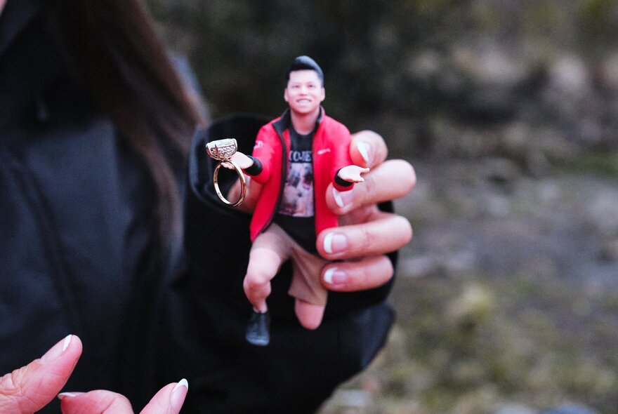 A realistic 3D printed figurine of a man down on one knee with palms outstretched, holding a real-life engagement ring; held by a woman.