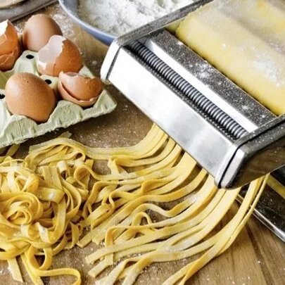 A stainless steel manual pasta machine rolling out fresh yellow noodles next to a carton of brown eggs and eggshells, and a bowl of flour.