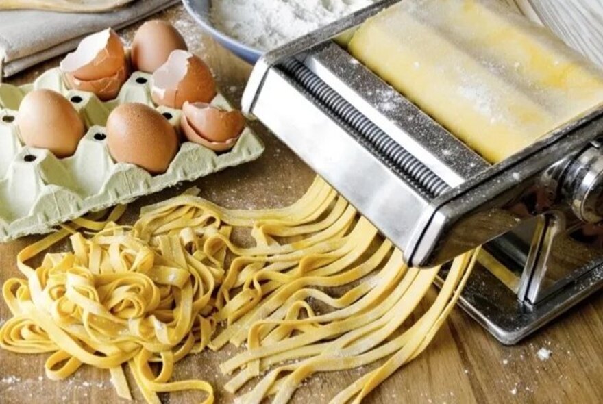 A stainless steel manual pasta machine rolling out fresh yellow noodles next to a carton of brown eggs and eggshells, and a bowl of flour.