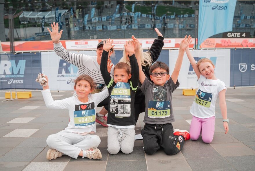 A group of kids kneeling and seated on the ground with their hands in the air at a running event. 