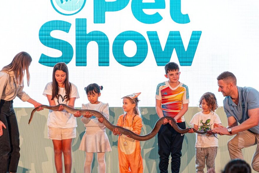 Five children standing in a line on a stage, using their hands to support a large snake, with an animal handler holding the snake's head; a partly obscured sign behind them group displaying the words PET SHOW.