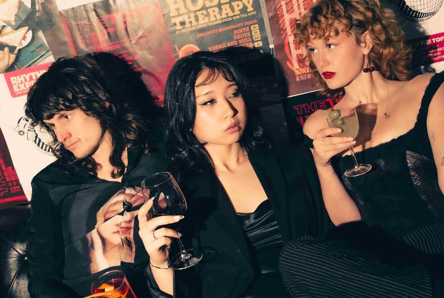 Three glamorous young people in black clothes drinking cocktails while leaning against a wall papered with band posters.