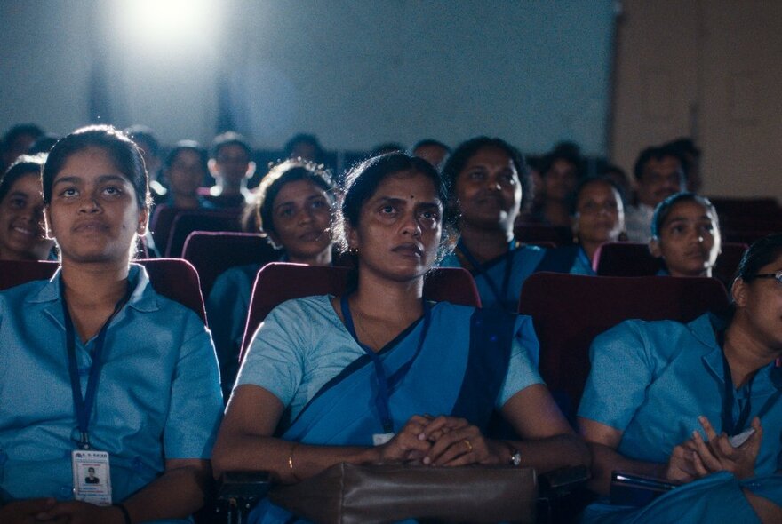 Film still of a seated audience of women in a cinema with the film projector lighting up the rear of the image, all of them wearing the same blue uniform and wearing lanyards around their necks.