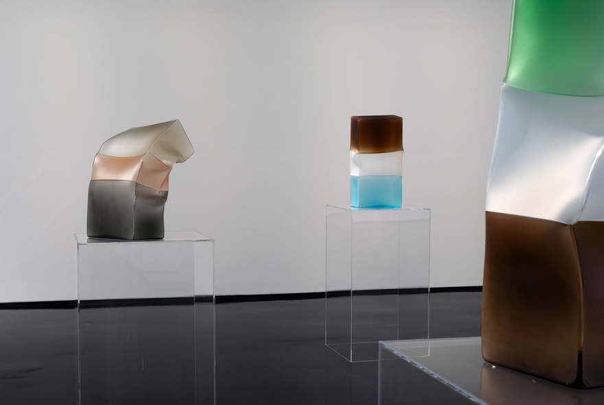 A gallery room featuring glass artworks made of three block colours, crumpled, on clear glass plinths.