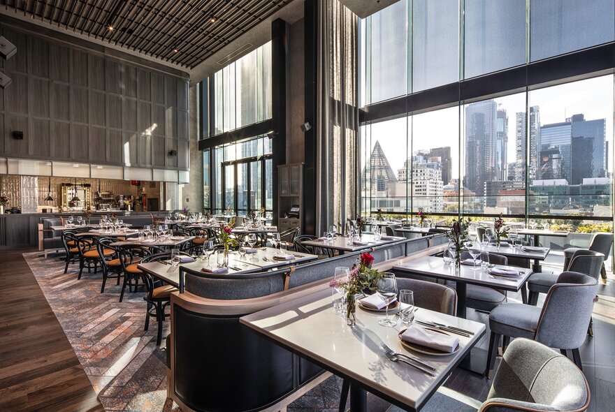 Modern restaurant interior featuring dining tables with white tops and grey upholstered chairs, large floor-to-ceiling windows looking out onto a city skyline.
