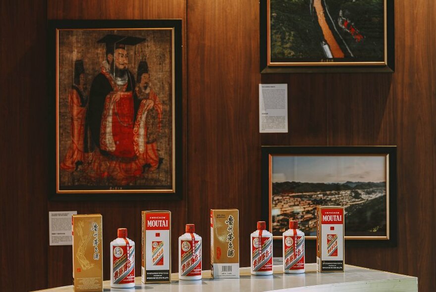 Row of bottles and boxes of bottles on a short white bar, three paintings on wooden wall behind.