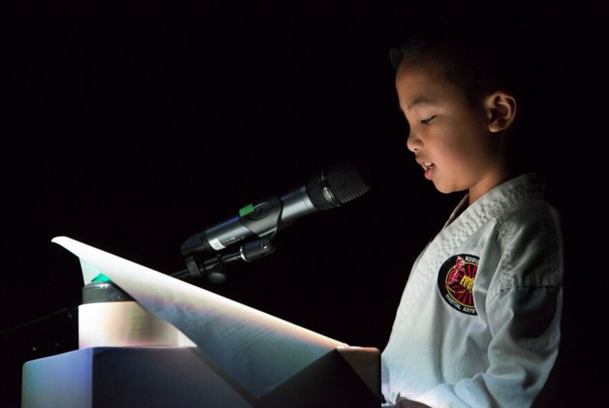 A child wearing a white kung fu outfit, reading from a piece of paper on a podium.