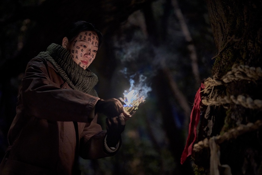 A scene from the film Exhuma showing a person in a dark forest at night, their face is covered in protective symbols and characters, and they are lighting a bundle of incense that glows and emits thick smoke.