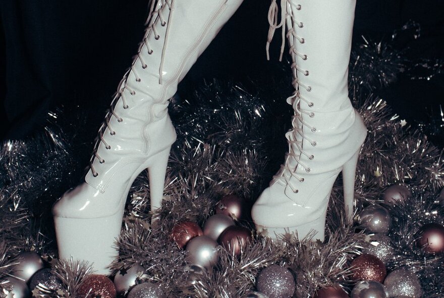 A close up of very high white platform boots standing on a carpet of Christmas baubles and tinsel. 