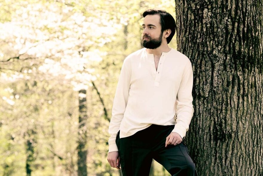 Bearded man wearing a white buttoned top, standing next to a large tree trunk in a forest with dappled foliage and light.