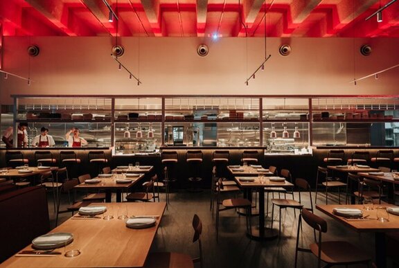 The empty interior of JUNI restaurant with tables set for service and two chefs in the kitchen.