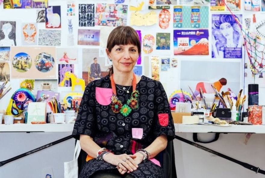 An artist shown in her studio with a work table and inspiration wall behind her.
