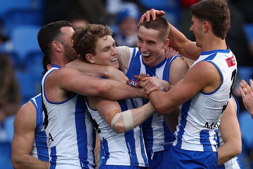 A group of North Melbourne AFL football players high-fiving and celebrating in a huddle on the field.