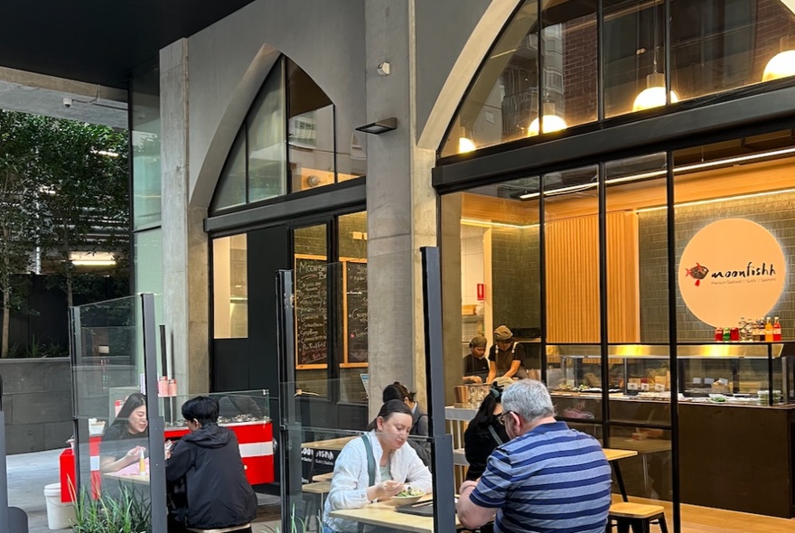 Exterior of the Moonfishh outlet showing people eating at outdoor tables, large glass windows, and a view into the shop.