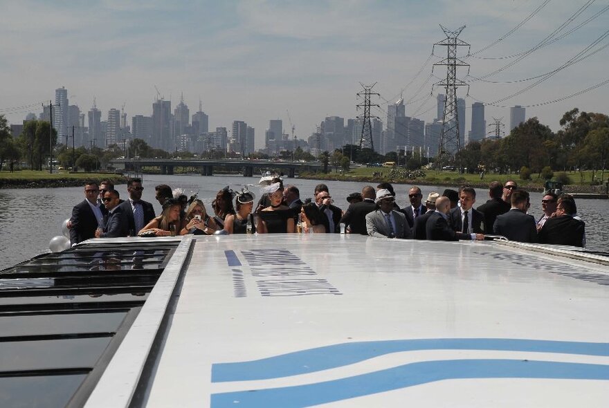 Crowd of well-dressed people standing at the back of a boat with the city skyline and river  behind them.