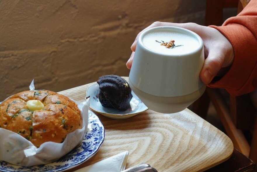 Hand holding drink in white pottery cup, with cake and madeleine on tray behind.