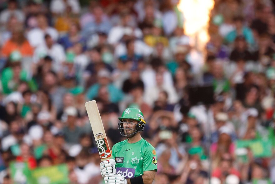 A Melbourne Stars BBL players holding his bat upwards with a blurred crowd in the background. 