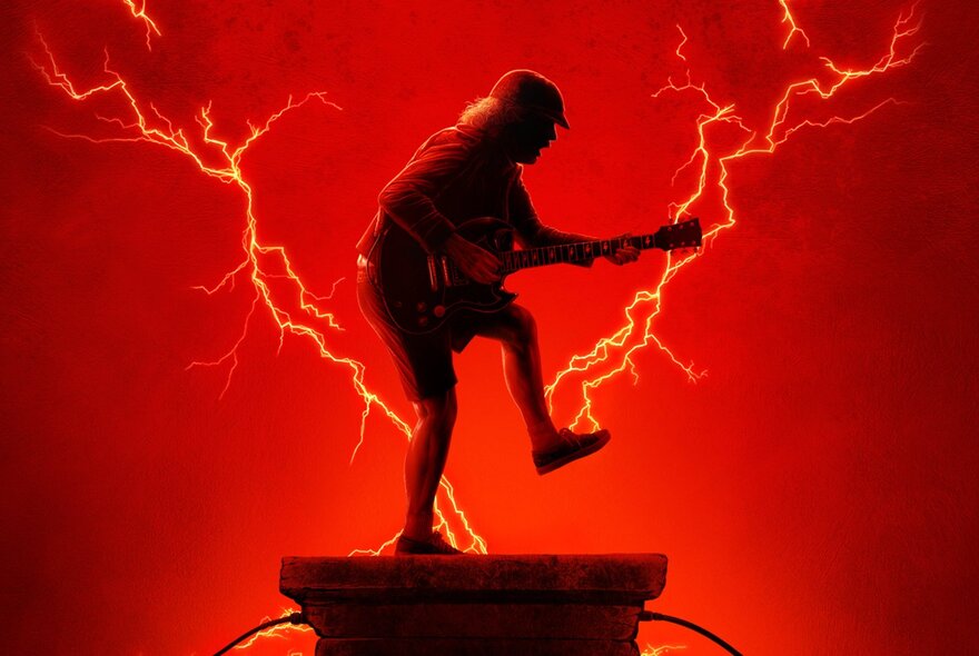 Lead guitarist Angus Young from the band AC/DC playing guitar, with one leg slightly raised off the ground, posed against a red background with flashes of lighting.