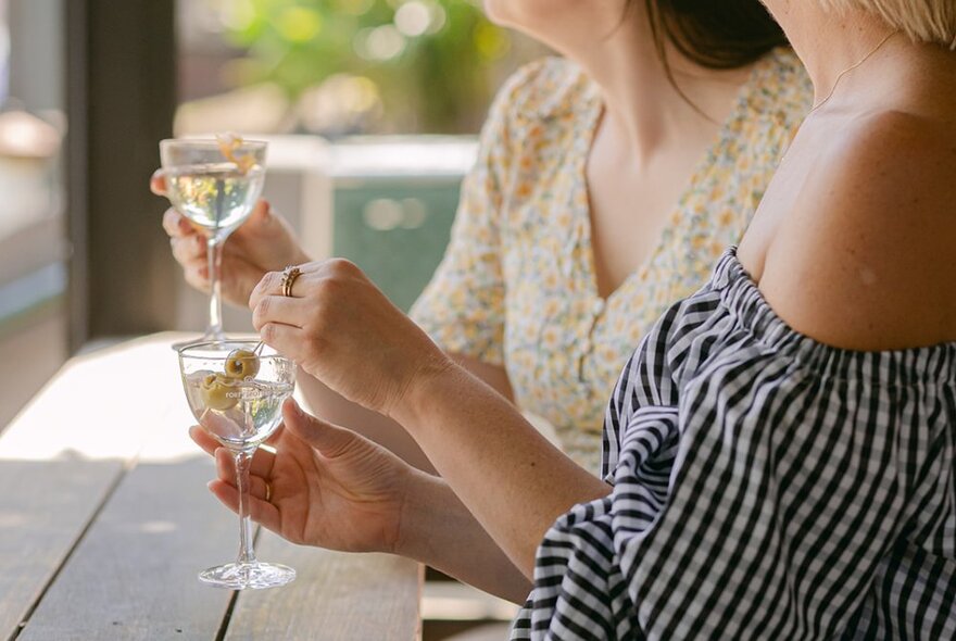 Two people sitting at an outdoor high table holding small martini glasses in their hands.