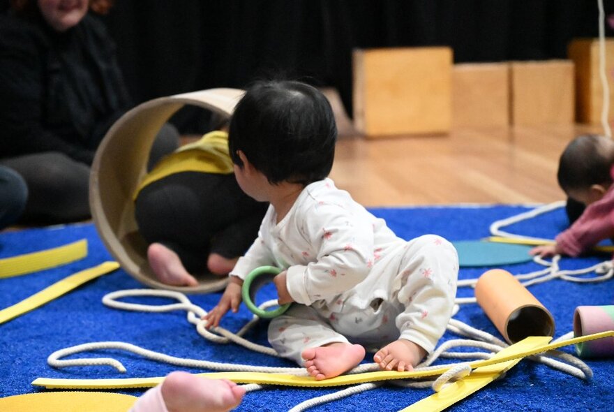 A baby in a play centre turning fully to view a another child crawling through a tunnel. 