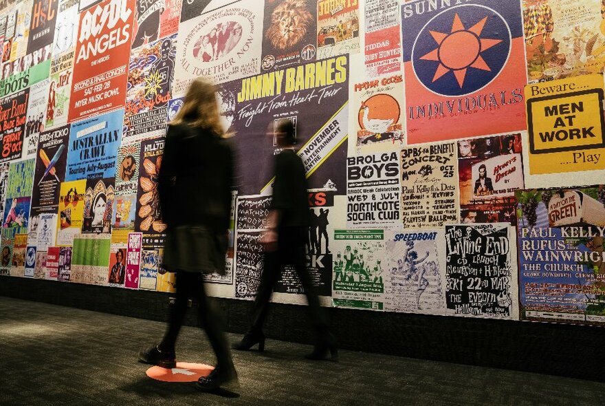 Two people, slightly blurred, walking along a wall of music posters.