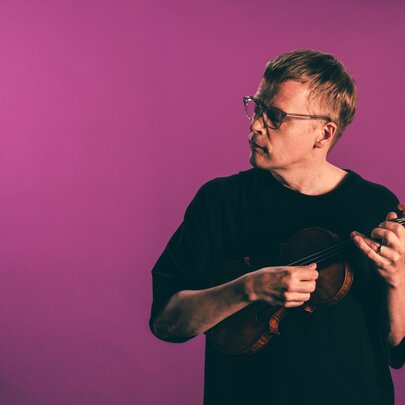 Finnish violinist and conductor Pekka Kuusisto holding a violin, posed against a magenta background.