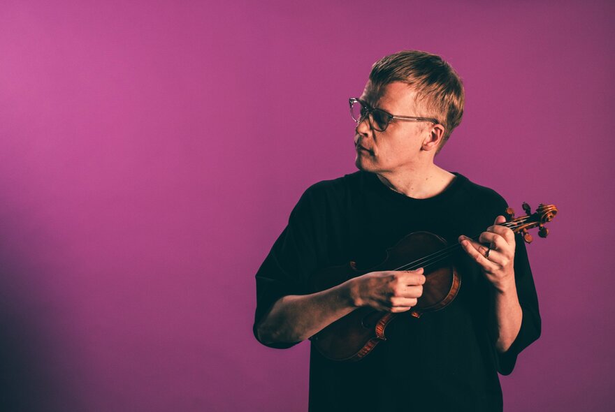 Finnish violinist and conductor Pekka Kuusisto holding a violin, posed against a magenta background.