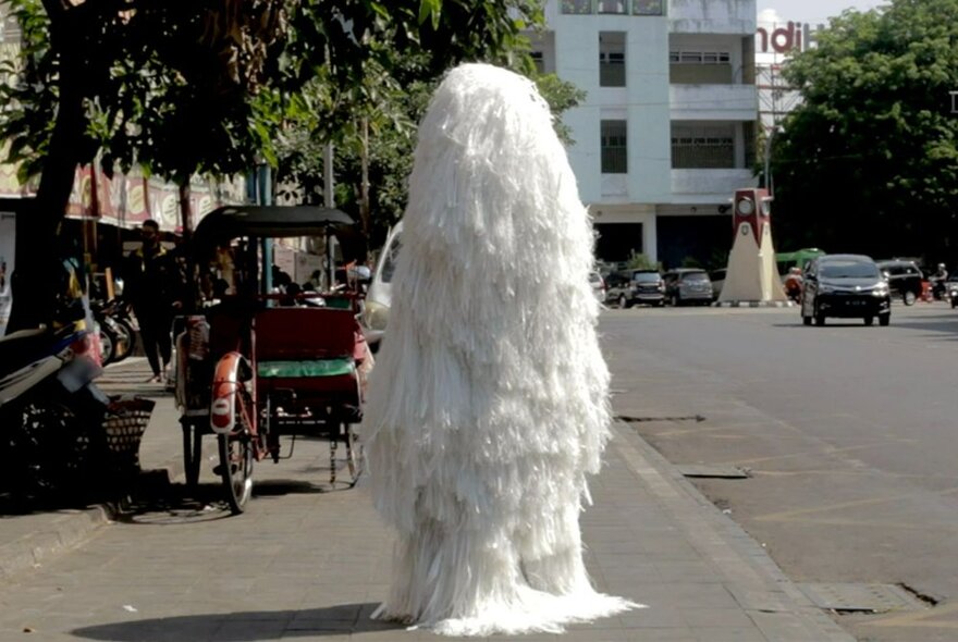 A large object made up of all white strands of feathers or shiny strips of white ribbon standing on a street in an Asian city in front of a bicycle rickshaw.