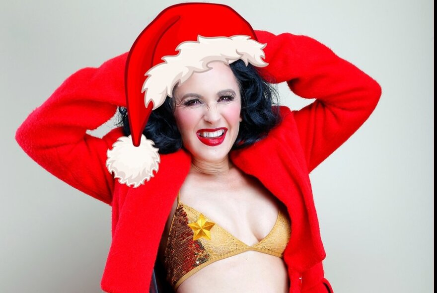 Smiling performer wearing a gold bikini top, an oversized red jacket and a red santa cap.